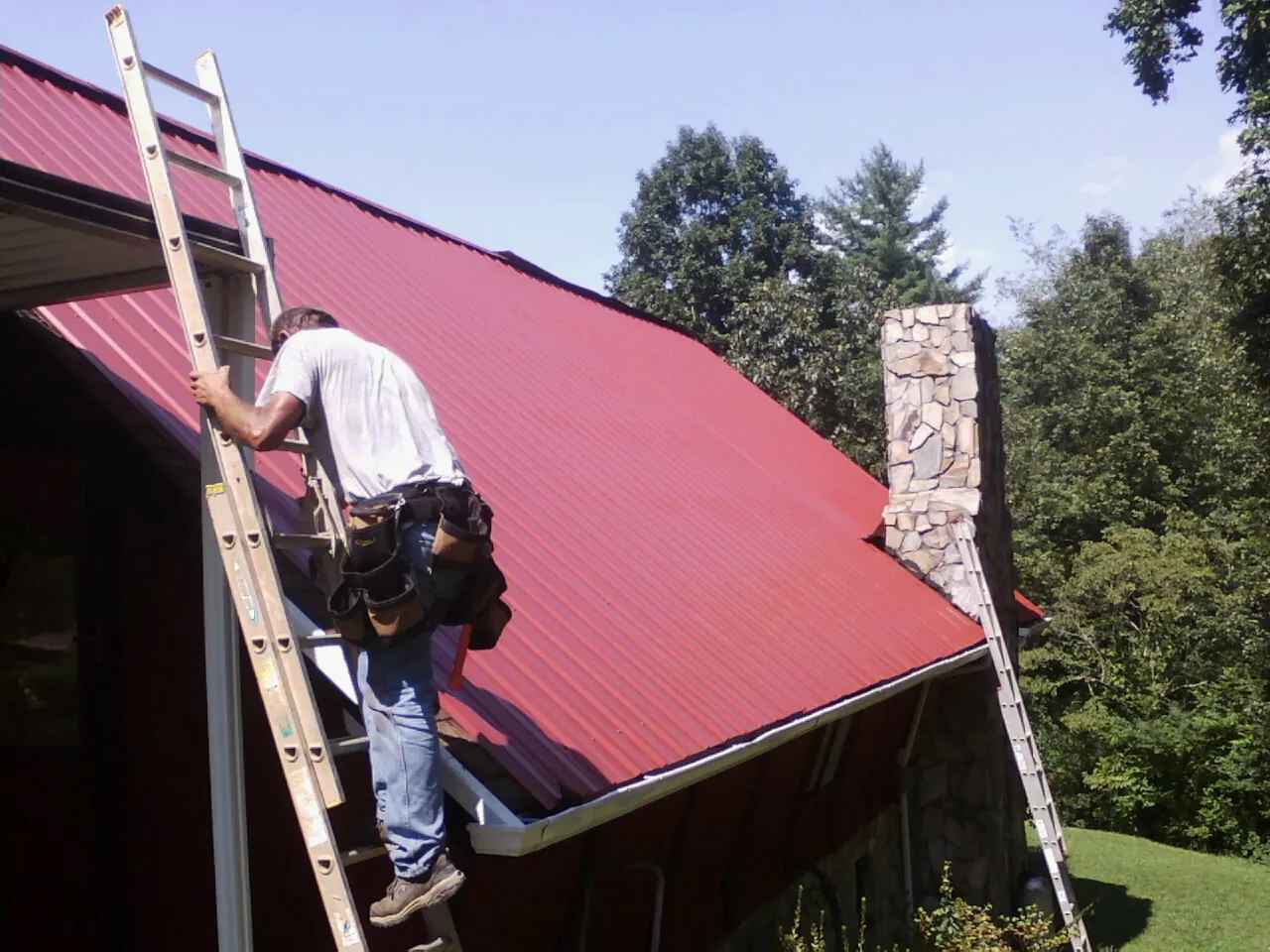 Roofer on a ladder beside a red metal roof and stone chimney — Soffit & Fascia Repair by Roofing Brothers in Hope